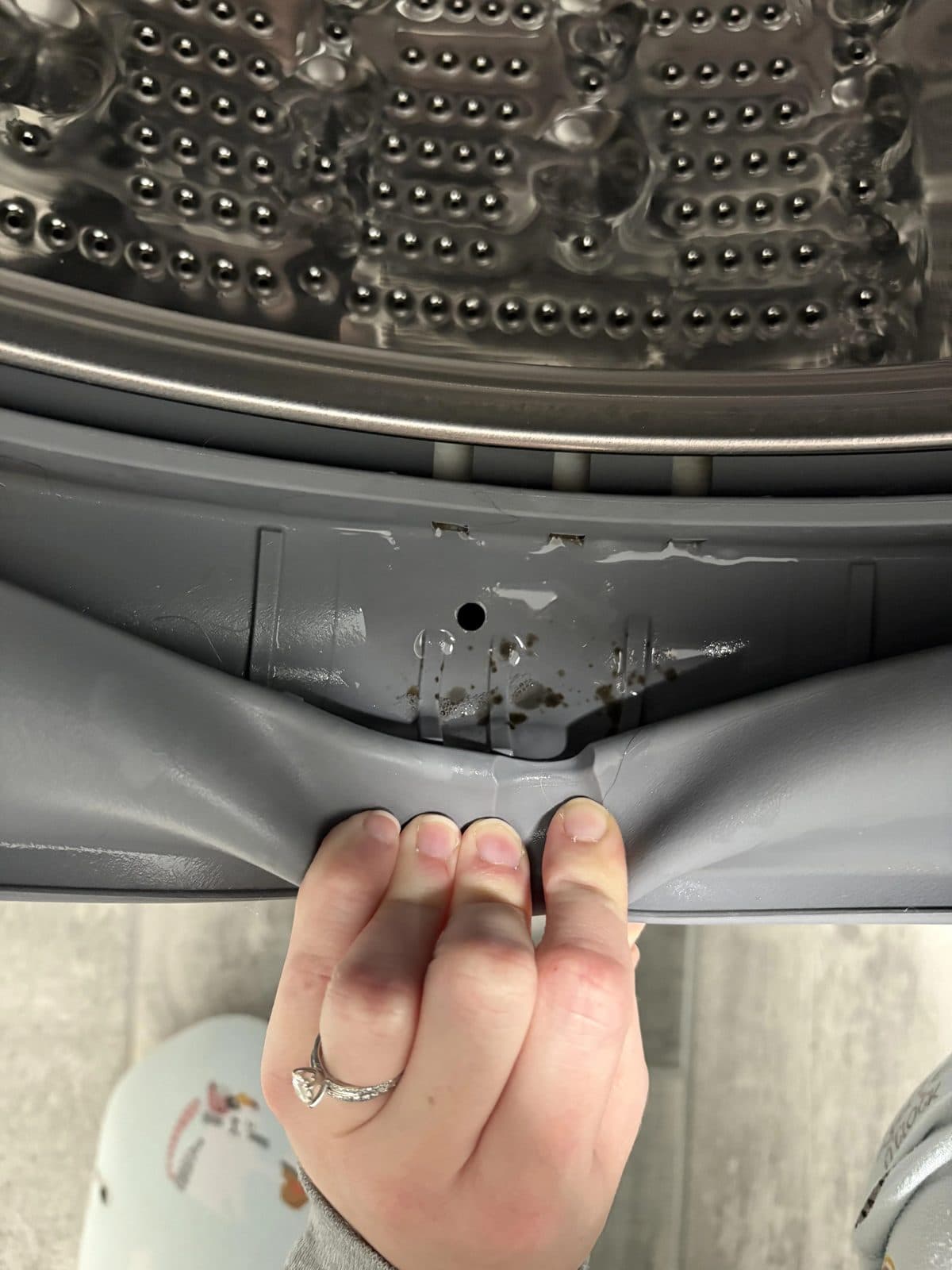 A front-load washer door gasket fold pulled back by hand, revealing trapped water and dark mold spots under the rubber lip